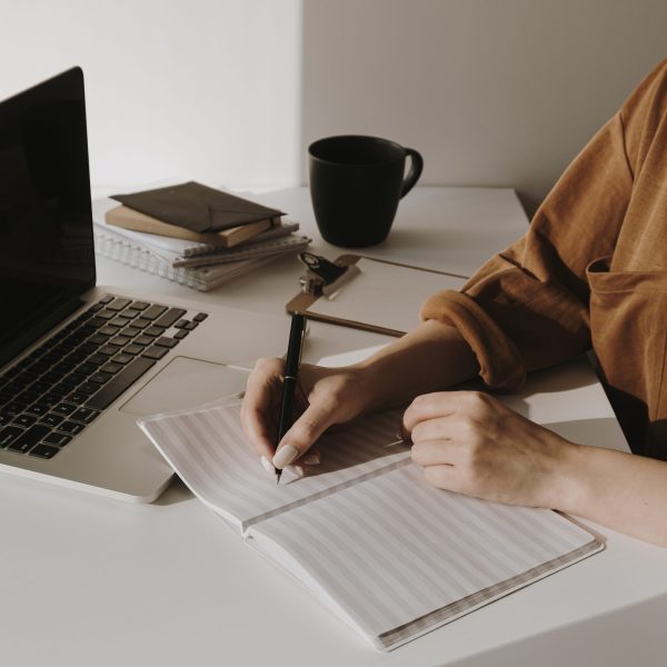 Minimalist home office workspace with laptop, coffee cup, clipboard. Woman write in a paper sheet notebook. Morning work, business concept. Sunlight shadows on the wall.